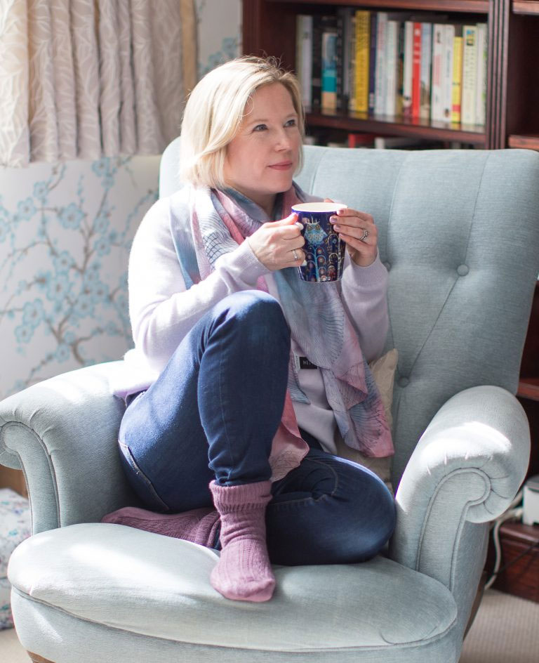 Professional audiobook narrator and voice coach, Esther Wane, sitting in a cozy armchair holding a mug, creating a warm and inviting creative studio atmosphere.